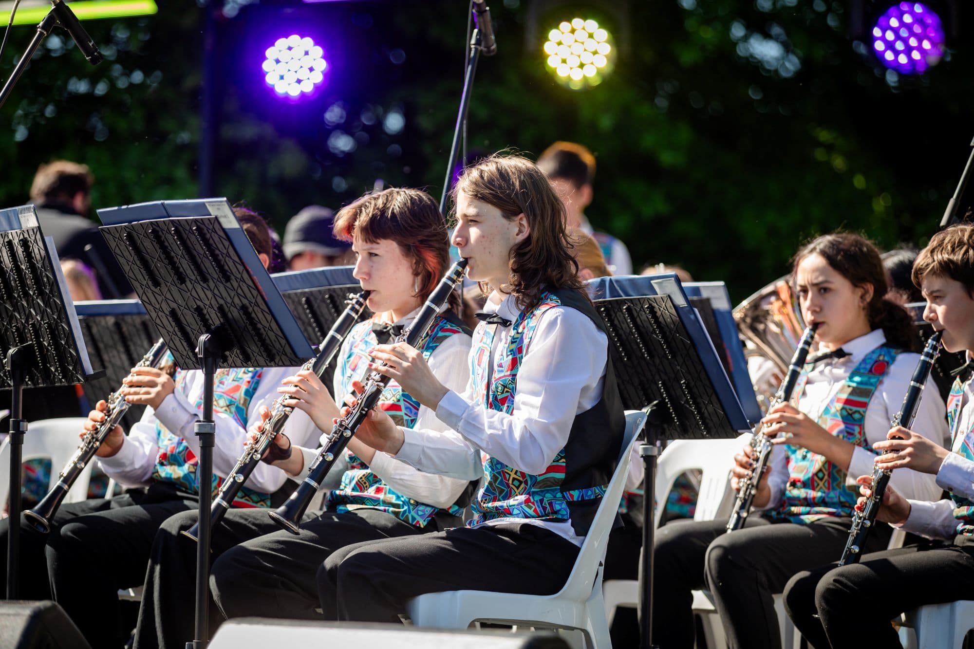 Members of a community concert band perform outdoors, seated with clarinets and sheet music stands beneath stage lighting.