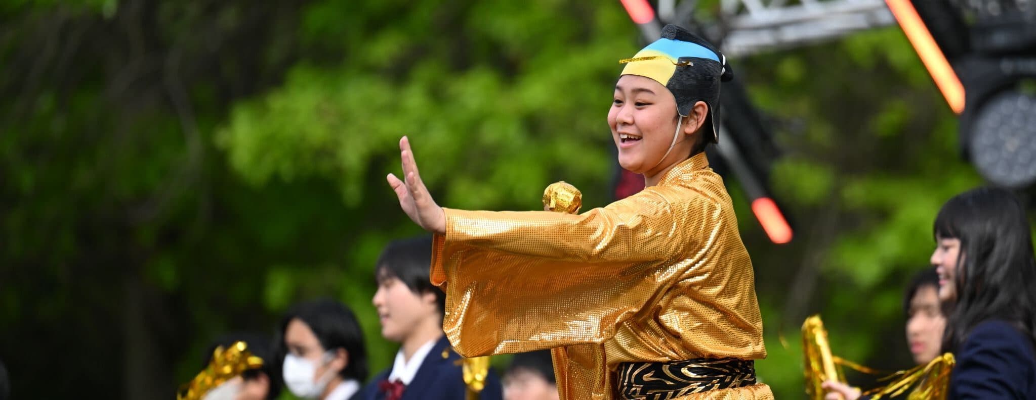 Stage performance featuring a dancer in a shimmering gold costume, accompanied by a group of performers holding gold pom-poms during an outdoor cultural event.